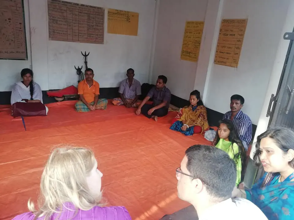 A group of adults and children sit on a red mat in a room with posters on the wall, listening to a discussion.