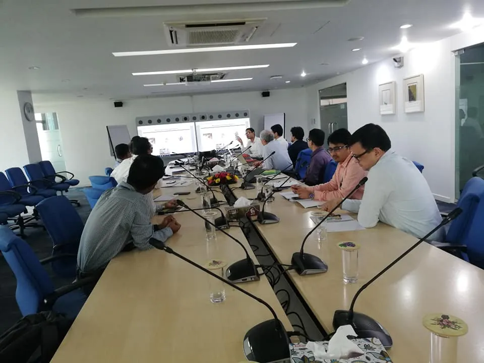 Wide view of a meeting room with a long conference table, desk microphones and two wall screens. Participants sit on both sides of the table.