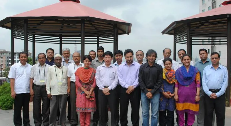 About twenty people, several wearing lanyards, pose for a group photograph between two garden pavilions with city buildings behind them.