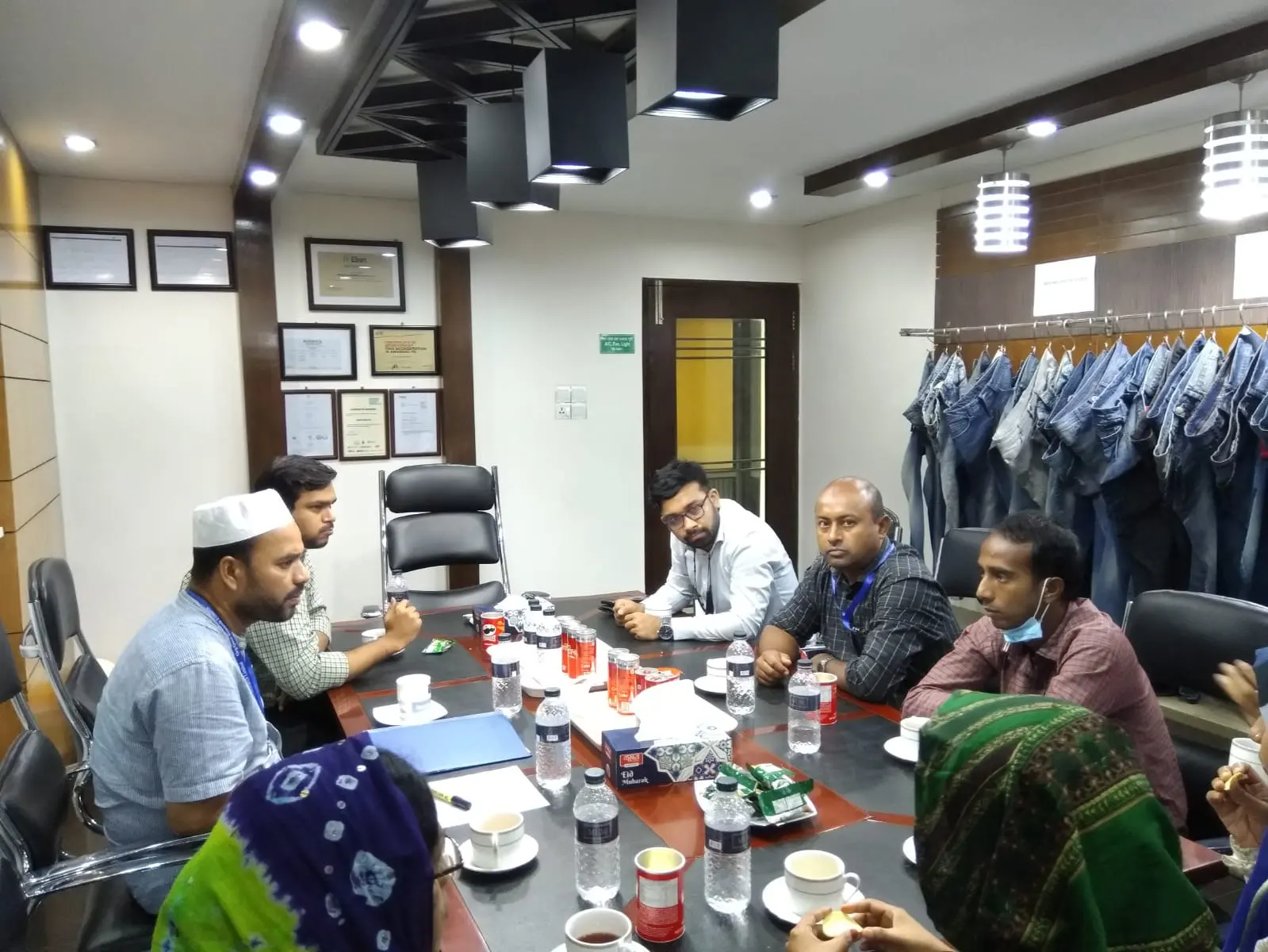 People sit around a table with cups and papers in an office. Denim garments hang on a rack behind them.
