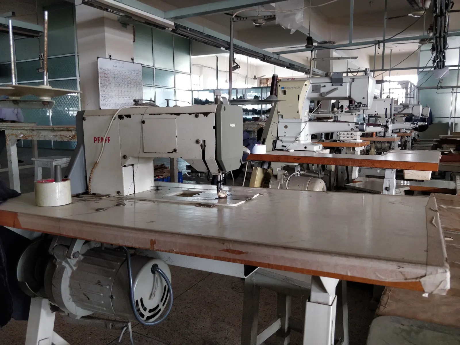 A long workshop table with industrial sewing machines in a row, and more machines and shelving behind.