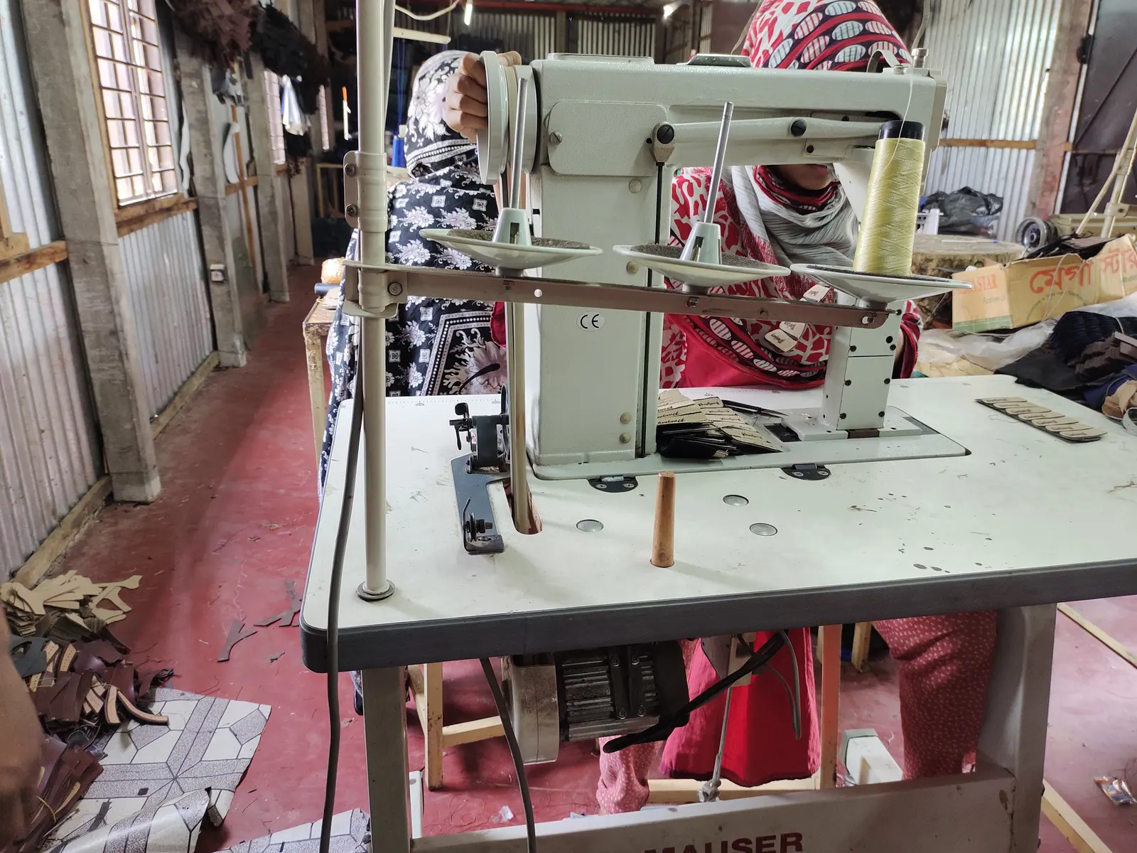An industrial sewing machine on a table in a workshop with corrugated walls; a worker is partly visible behind it.