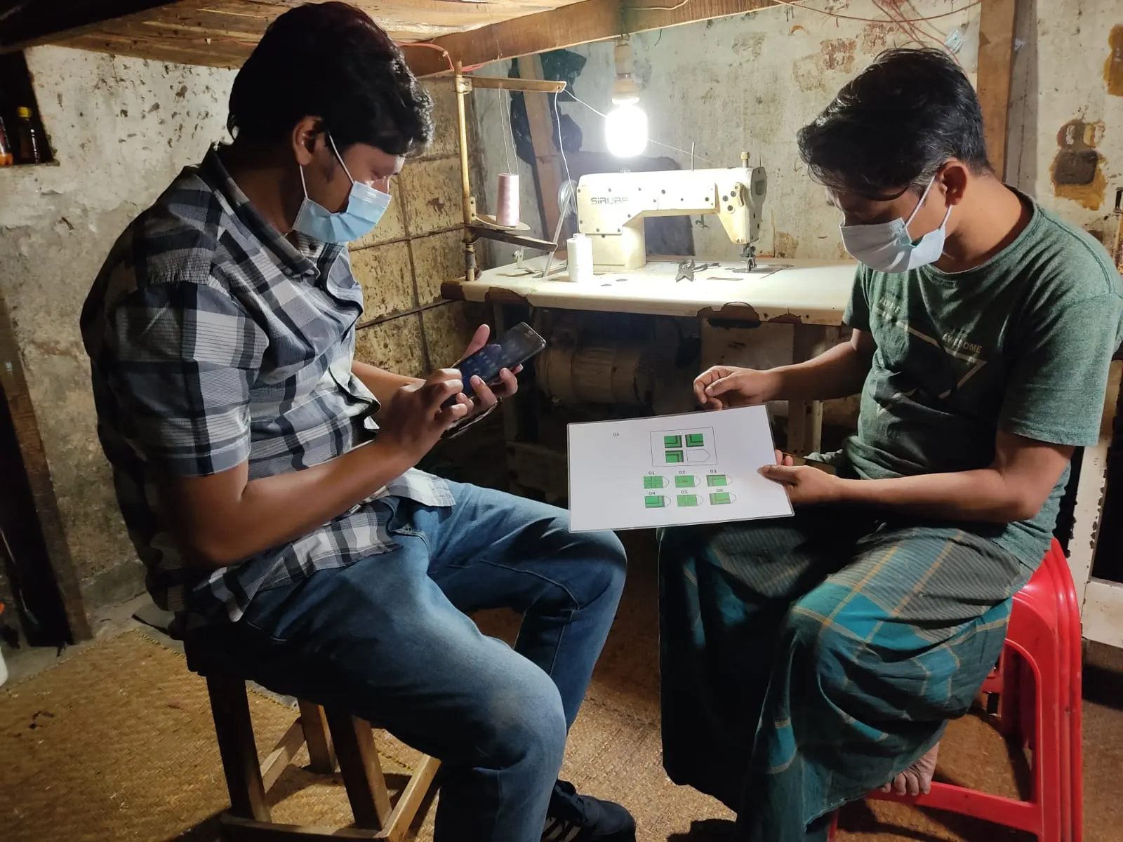 Two men wearing face masks sit in a workshop. One holds a phone; the other holds up a sheet printed with a green diagram.