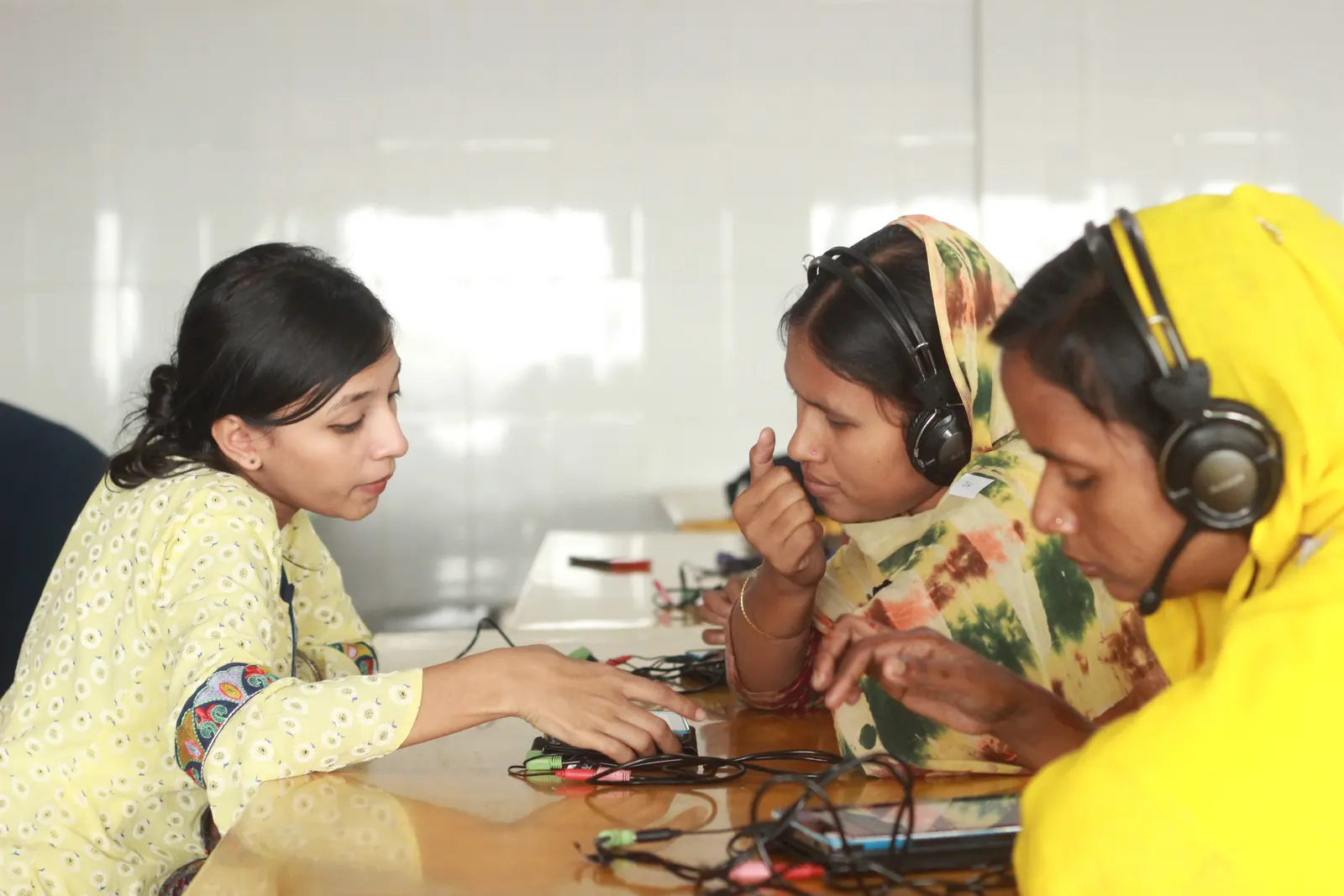 A young woman helps two women in headscarves, who wear headphones, with a tablet at a table.