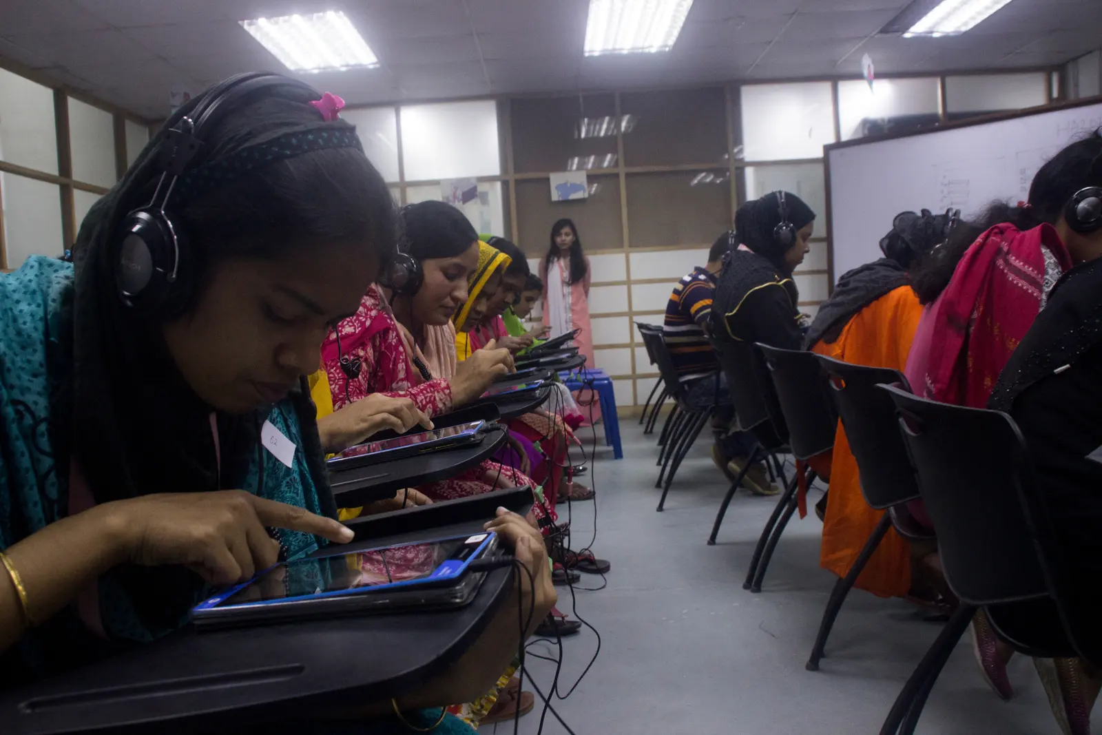 Women wearing headphones tap tablets at a row of desks in a classroom-style room.