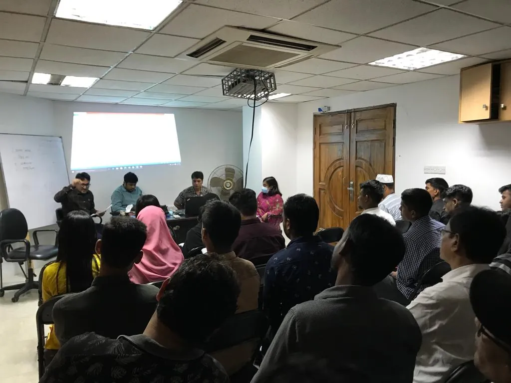 Rows of seated participants face a front desk where trainers sit under a projected screen, in a small training room with a whiteboard.