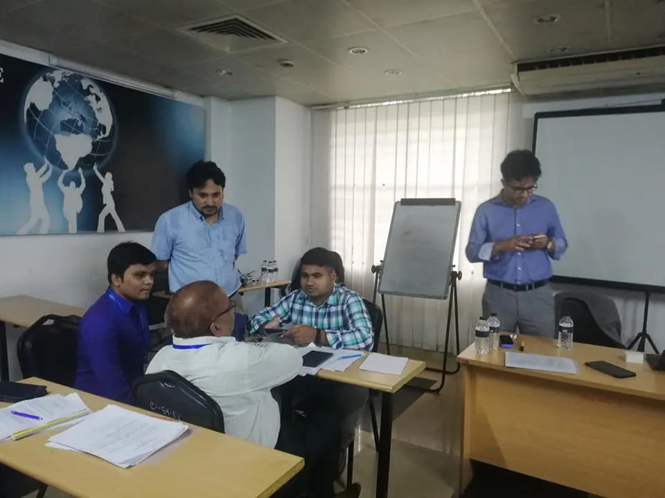 Participants at desks with papers in a training room, with a flip chart behind them. One man stands behind the seated group and another stands at the right using a phone.