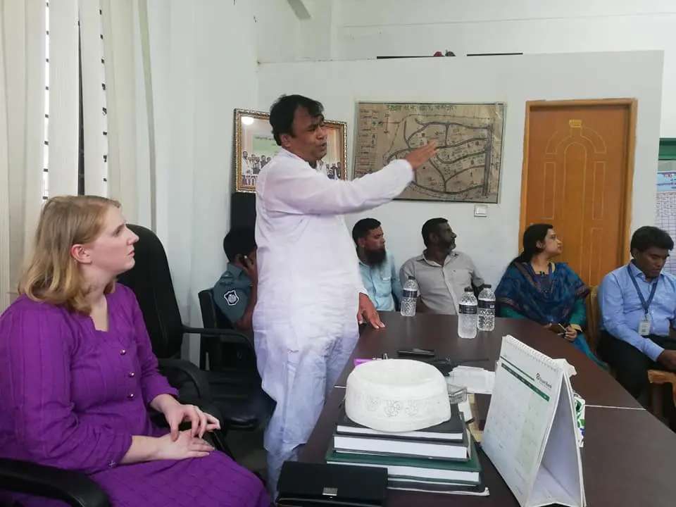 A man in white stands and points at a framed wall map while people sit around a table.