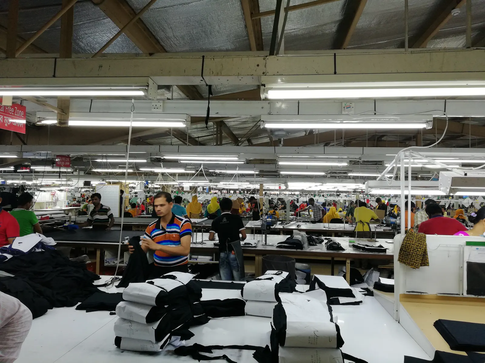 A garment factory floor with long rows of cutting and sewing tables under strip lights; workers stand at the tables and a man in a striped shirt is in the foreground.