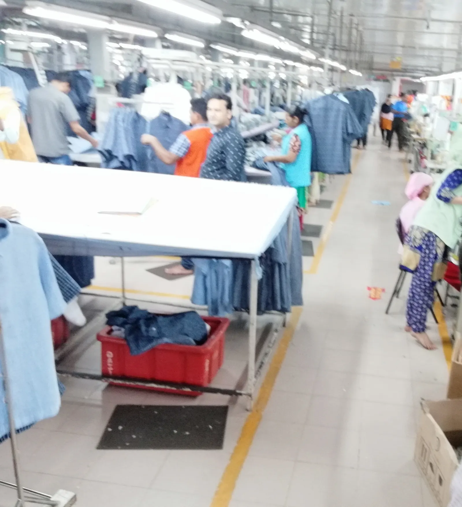 A production aisle in a garment factory: a white table and a red crate in the foreground, workers and hanging denim garments along the line behind.