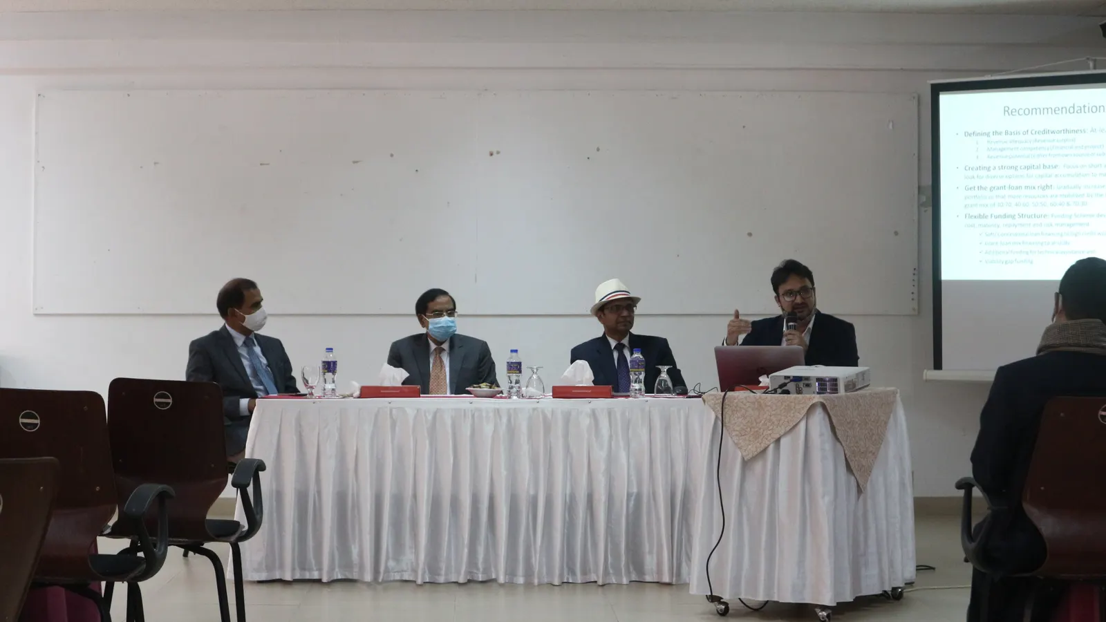 Four speakers, some wearing face masks, sit at a white-draped head table beside a projected slide in a seminar room.