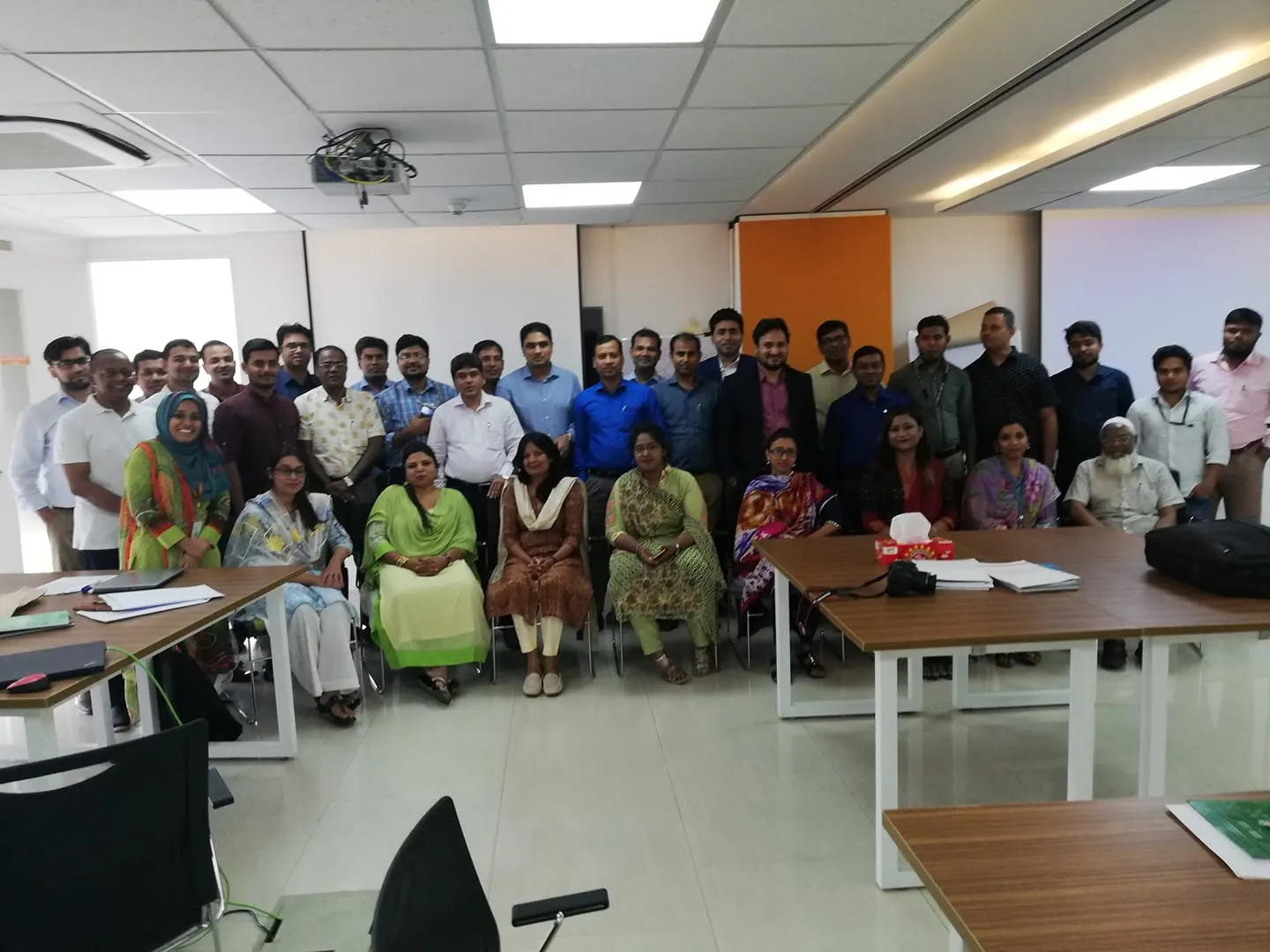 About forty people pose for a group photograph in a modern training room with desks and a projector.