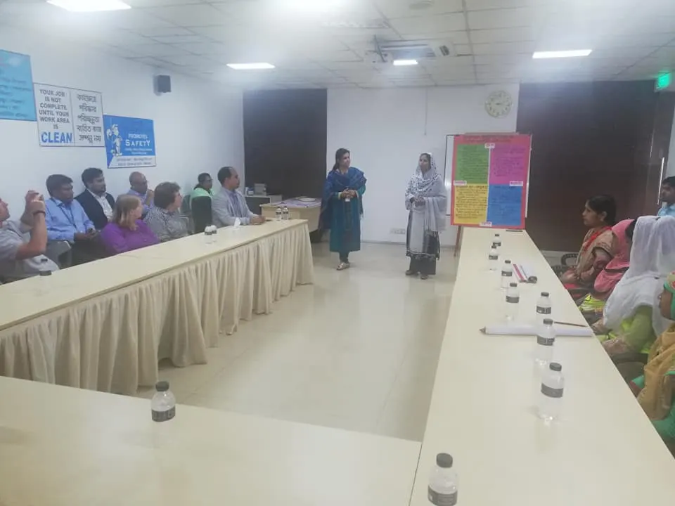 A workshop in a long meeting room; two women stand by a flip chart at the end of the table and posters about hygiene hang on the wall.