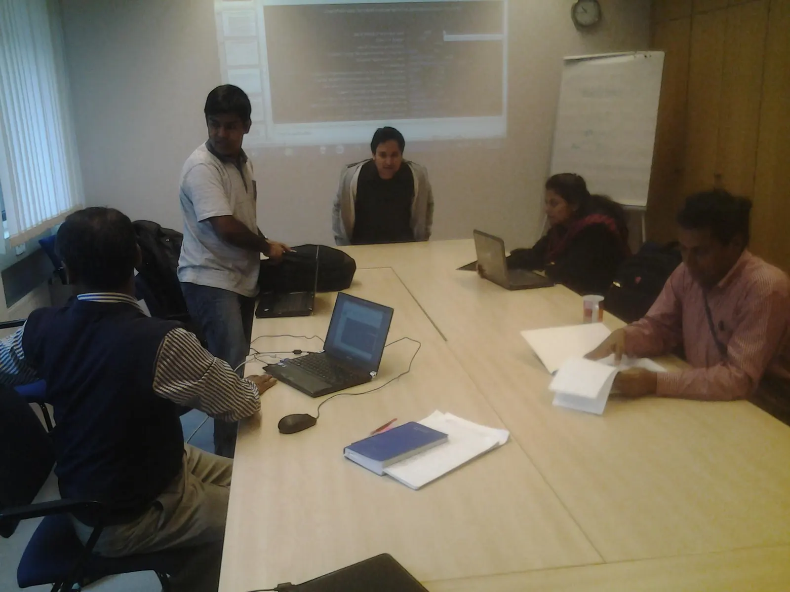 People work with laptops and papers around a meeting-room table, with a projection screen and a flip chart behind them.