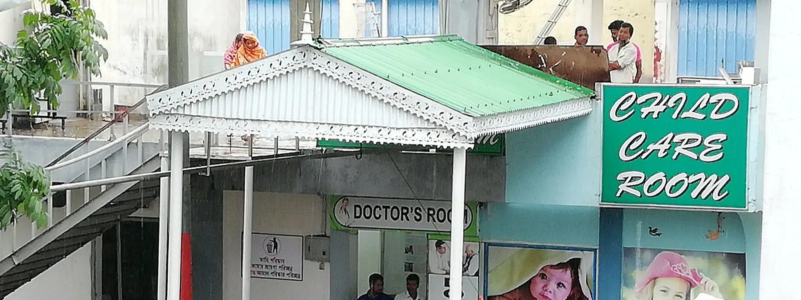 A single-storey building with a green roof and signs reading ‘Doctor’s room’ and ‘Child care room’, beside an open stairway.